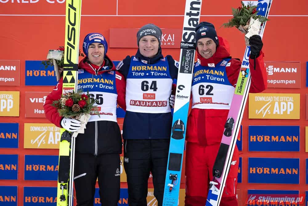 Trzech uśmiechniętych skoczków narciarskich na podium. Trzymają w ręce bukiet kwiatów